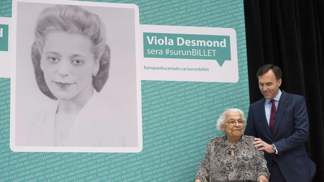 Le ministre des Finances, Bill Morneau, pose une main sur l'épaule de Wanda Robson, soeur de Viola Desmond, devant une affiche annonçant que Desmond a été choisie pour figurer sur les billets de 10 $.