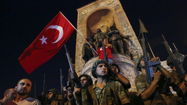 Un homme tient un drapeau turc devant le monument de la république, derrière les soldats armés.