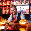 Un homme portant une chemise et une cravate se trouve derrière le bar et sourit. De nombreuses bouteilles d'alcool se trouvent derrière lui et il est entouré de cocktails, de citrouilles et de gens célébrant l'Halloween.