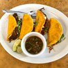 Four tacos on a plate with lime wedges and a small bowl of broth.
