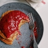 Deux crêpes empilées et garnies de compote fraises et dattes déjà entamées à côté d'une fourchette, dans une assiette, et un contenant de compote.