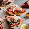 Des bouchées de tartines aux tomates, aux olives et au prosciutto réparties sur une planche, accompagnées d'un quartier de citron.
