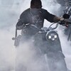 Un homme avec des lunettes fumées, une tuque ornée de flames et une cigarette entre les lèvres conduit une motocyclette, entouré de fumée.