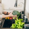 Une personne lave du brocoli et d'autres légumes sous un jet d'eau au-dessus d'un évier.