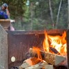 Un feu de camp brûle dans un foyer de métal avec, en arrière-plan, un campeur assis à une table à pique-nique.