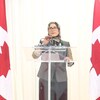Amira Elghawaby en conférence de presse parle au micro, encadrée par deux drapeaux du Canada.