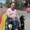 Anaïs Sabourin se déplace en fauteuil roulant en compagnie de sa chienne d'assistance, Mika. Ils sont sur la rue Ste-Catherine. Des arc-en-ciel ont été dessinés sur la rue.