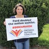 Angela Brandt, debout à l'extérieur, tenant une affiche sur laquelle il est inscrit en anglais "Ford doubled the autism waitlist. 50K is not OK". 