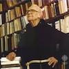 Le père Anselme Chiasson appuyé sur une pile de livres dans une bibliothèque. 