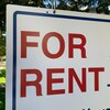 Un passant marche sur le trottoir et croise une affiche sur laquelle les mots « For Rent » sont écrits en rouge.