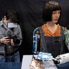 Le robot à l'apparence de femme peint sur un canevas posé sur une table à l'aide d'un bras mécanique. Une personne derrière le robot le photographie.
