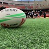Un ballon de rugby en avant-plan sur le gazon artificiel du terrain du stade BC Place. Un groupe de joueurs de rugby en arrière-plan.