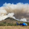 Des panaches de fumée du feu de forêt Battleship Mountain derrière une montagne et un hélicoptère qui est posé sur un champs.