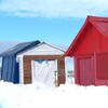 Trois cabanes en bois dans la neige, sur le bord de l'eau. Celle à l'avant-plan est peinte en rouge. Une est peinte en blanc et une autre est bleue avec une étoile jaune pour représenter le drapeau acadien.