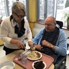 Une femme debout fait manger un homme âgé assis dans une chaise roulante devant un plateau de nourriture.