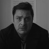 Un homme à l'air inquiet regarde droit devant lui, alors qu'il est assis dans une salle de bains, dans une image en noir et blanc de la série Bon matin Chuck.