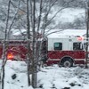 Un camion de pompiers en mouvement, ses lumières d'urgence allumées, est photographié entre des branches d'arbres sur une route rurale enneigée.