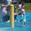 Un père à genoux dans l'eau replace le bonnet d'un petit enfant, debout dans les jeux aquatiques d'un parc, en été.