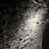 Un mur de pierre dans un couloir souterrain est illuminé partiellement par la lampe de poche que tient une personne.