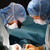 Dans une salle d'opération, deux personnes portant des combinaisons bleues et des masques chirurgicaux sont face à face, et sont penchées au-dessus d'une table d'opération où est couché un patient, hors du cadre de la photo.