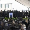 Un groupe de personnes habillées en noir chantent sur une scène surélevée. Devant elles, une foule, également vêtue de noir, les écoute.
