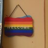 Au premier plan, une affiche aux couleurs de l'arc-en-ciel, posée sur le mur, indique "Welcome". À l'arrière-plan, une femme assise à une table parle au téléphone.