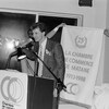 Photo en noir et blanc. François Rioux parle à un micro. Derrière, deux hommes tiennent un drapeau sur lequel il est écrit « soixante-quinzième anniversaire, La Chambre de commerce de Matane, 1913-1988. »