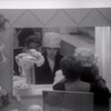 Une femme essaie un chapeau devant un miroir pendant qu'une vendeuse assise derrière elle procède aux derniers ajustements.