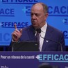 Le ministre de la Santé, Christian Dubé, parle au micro devant un mur bleu sur lequel apparaît plusieurs fois la mention « Plus efficace ».