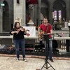 Un homme joue de la guitare et une femme du violon. Ils sont à l'intérieur de la cathédrale de Monc à Moncton. 