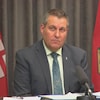 Le ministre Cliff Cullen est assis devant un rideau gris et deux drapeaux du Manitoba et il s'apprête à parler dans un microphone placé sur une table.