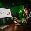 Une personne concentrée regarde son ordinateur portable et un plus gros écran sur son bureau dans un décor sombre avec des lumières vertes. 