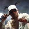Un homme portant une veste de sport blanche, une casquette blanche et des lunettes fumées tient un micro dans la main droite. 
