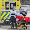 Deux personnes poussent une civière sur laquelle se trouve une patiente, près d'une ambulance.