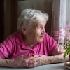 Une dame âgée est assise à une table et regarde par la fenêtre.