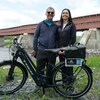 Un homme et une femme derrière un vélo électrique. À l'arrière-plan on peut apercevoir un long pont couvert rouge.