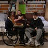 Deux personnes en fauteuil roulant mangent autour d’une table dans une chambre d’hôpital. Une troisième personne lit un livre.