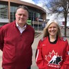 Mike Savage porte un gilet rouge et Dawn Arnold porte un chandail de hockey d'Équipe Canada. Ils posent tous les deux dehors devant un aréna.
