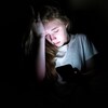 The image displays an upset girl sitting in the dark while using her smartphone. The light from the screen is illuminating her face.