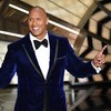 Dwayne Johnson porte un costume en velours bleu et il pointe les doigts vers le ciel.