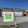 Photo extérieure d'une école, un bâtiment de deux étages. À l'avant-plan, une affiche de félicitations pour un projet de cour d'école accrochée sur une clôture.