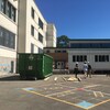 Une cour d'école avec des lignes colorées au sol; un conteneur vert, une école modulaire en chantier et trois employés portant des casques de construction par une journée ensoleillée