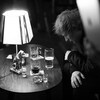 L'artiste est assis devant une table remplie de verres de bière et d'alcool; il regarde le sol, les bras croisés. 