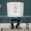 Un carton identifié par le logo d'Élections Nouveau-Brunswick cache le visage d'un électeur assis, dont on ne voit que les jambes.