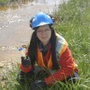 Nadia Mykytczuk porte un casque et des gants près d'un plan d'eau. Elle tient une éprouvette remplie d'eau dans une main.