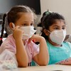 Deux petites filles d'environ 6 ans sont assises à leur pupitre dans une salle de classe. Elles portent des masques N95. 