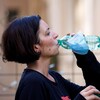 Une femme photographiée de profil dans une salle d'entraînement. Elle prend une gorgée d'eau d'une bouteille autour de laquelle est enroulé un masque médical bleu.