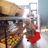 Une femme qui tient un petit panier d'épicerie s'accroupit pour prendre une pomme dans une caisse posée sur la rangée du bas dans une épicerie de quartier.

