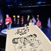 Une scène de théâtre avec des acteurs et un programme en premier plan avec le titre "C'est à nous de voir".
