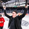 Sur la glace d'un aréna, un jeune homme soulève, victorieux, un grand trophée.